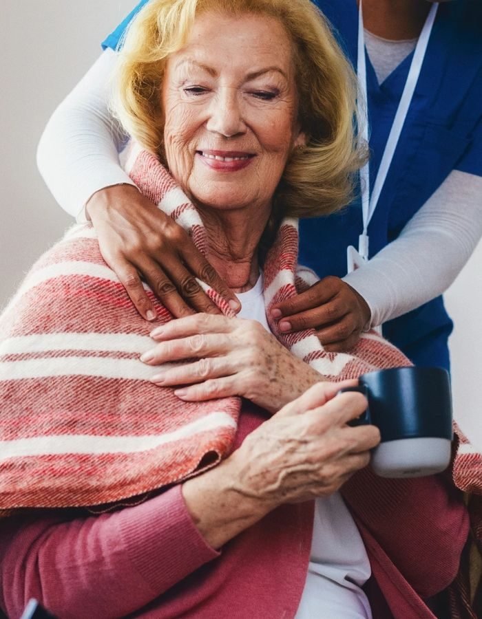 A smiling older woman wrapped in a blanket holding a mug while a caregiver rests reassuring hands on her shoulders